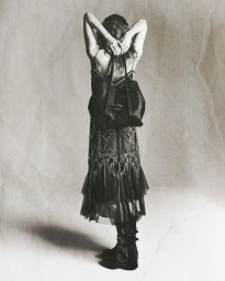 Model standing with back to camera wearing embroidered slip dress, boots, and holding handbag against textured backdrop
