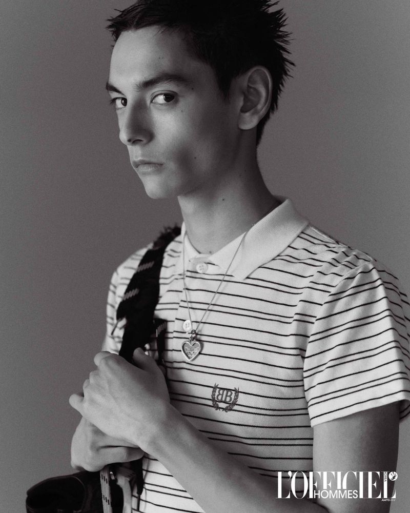 Monochrome portrait of a young adult man with short hair wearing a striped collared shirt and a heart pendant necklace.