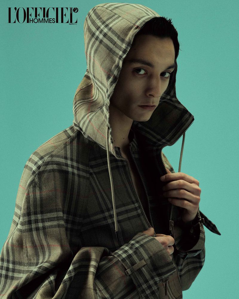 Model wearing a plaid hooded jacket adjusting the zipper against a teal background for L'Officiel Hommes Korea.