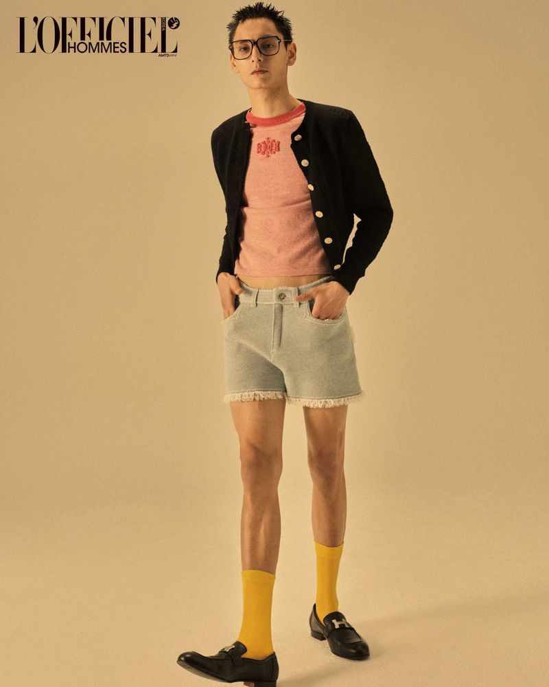 Fashion editorial shot of a young man in a black cardigan, pink t-shirt, and denim shorts, paired with yellow socks and black loafers.