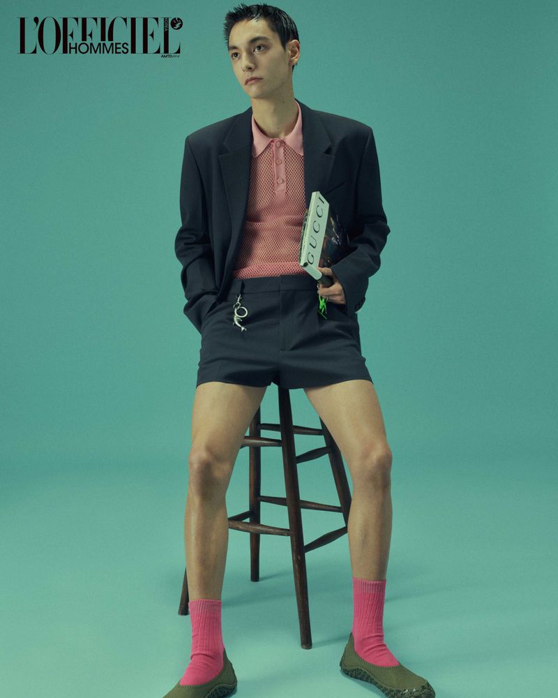 A young adult male model poses in a studio wearing a black suit jacket, shorts, pink socks, and green shoes while holding a Gucci book.