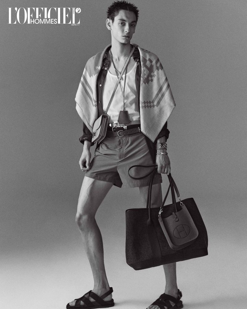 Monochrome portrait of a male model wearing shorts, sandals, shirt and carrying bags in a studio setting.