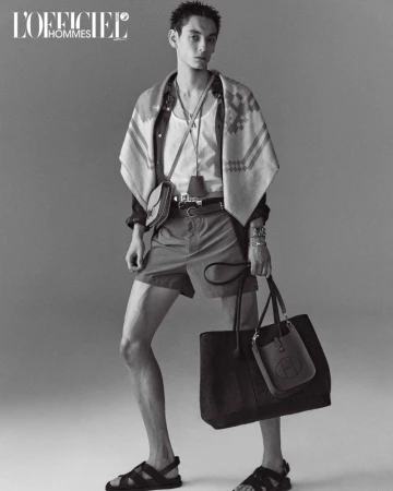 Monochrome portrait of a male model wearing shorts, sandals, shirt and carrying bags in a studio setting.