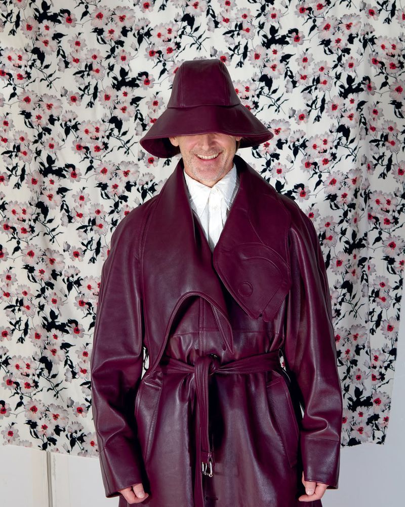 Smiling man wearing a burgundy leather hat and belted trench coat with a floral pattern backdrop, eye-level angle, indoors.