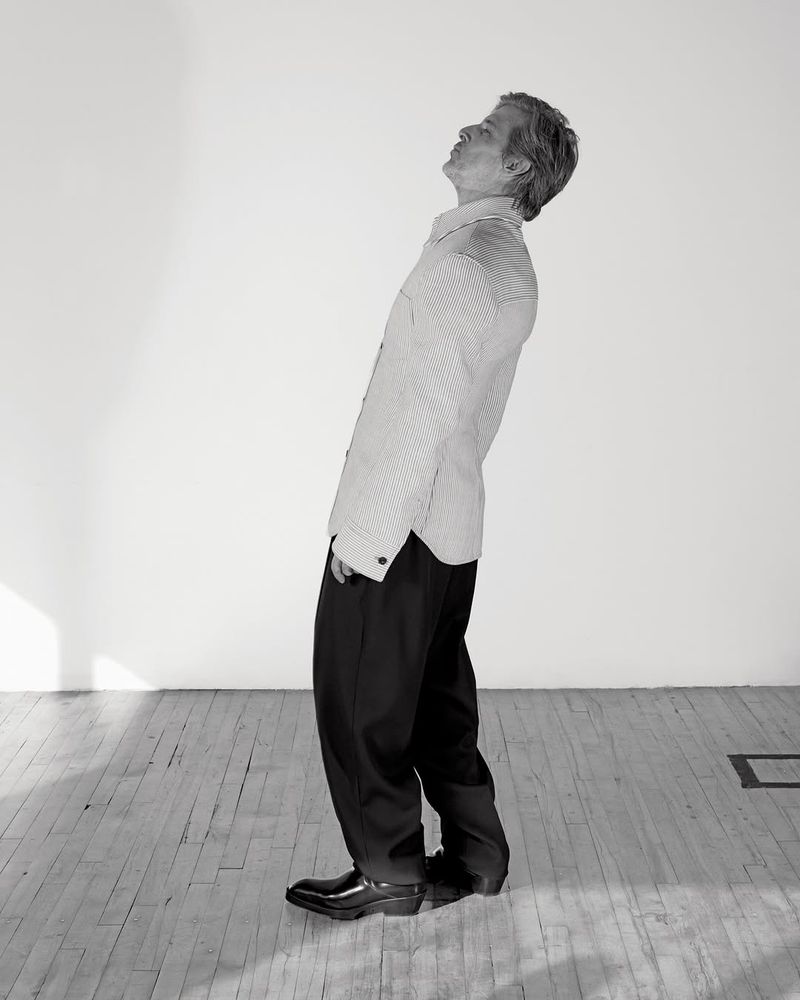 Full shot of an adult male in a white striped shirt and black pants, leaning back in a photo studio against a plain white background.