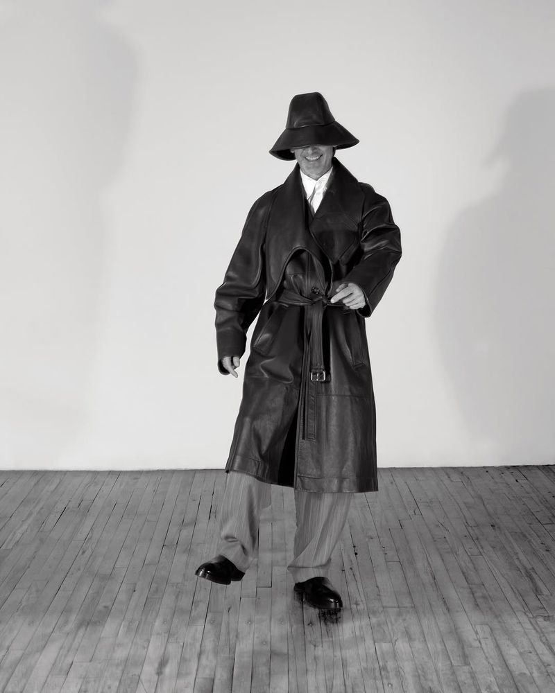 Monochrome portrait of an adult wearing a wide-brimmed hat and leather trench coat, set against a plain backdrop and wooden floor.