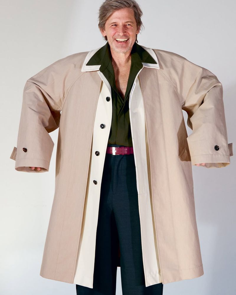 Portrait of an adult man smiling, wearing an oversized tan trench coat with an olive-green shirt and dark pants, standing in a white studio.