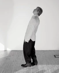 Man leaning backwards wearing a patterned shirt and dark trousers in a room with a wooden floor and plain white walls