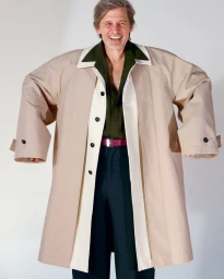 Man wearing an oversized beige trench coat with exaggerated wide shoulders standing against a white seamless background