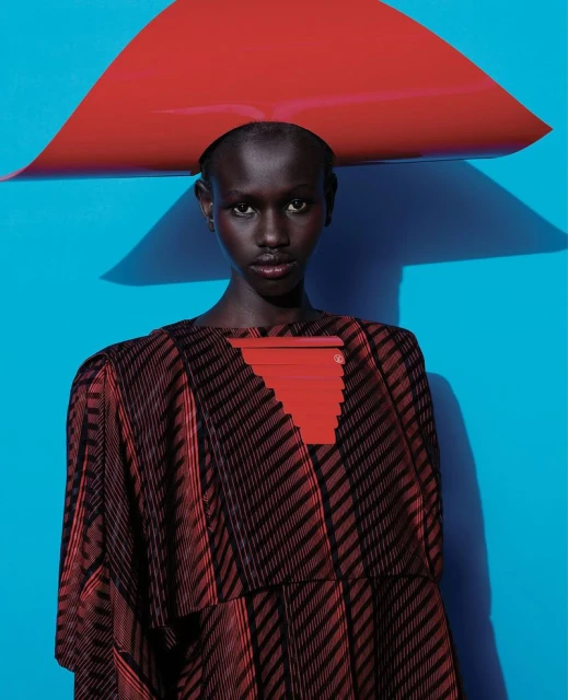 Fashion-forward portrait of a Black woman in a striped dress and red headpiece against a bright blue backdrop, highlighting bold colors and style.