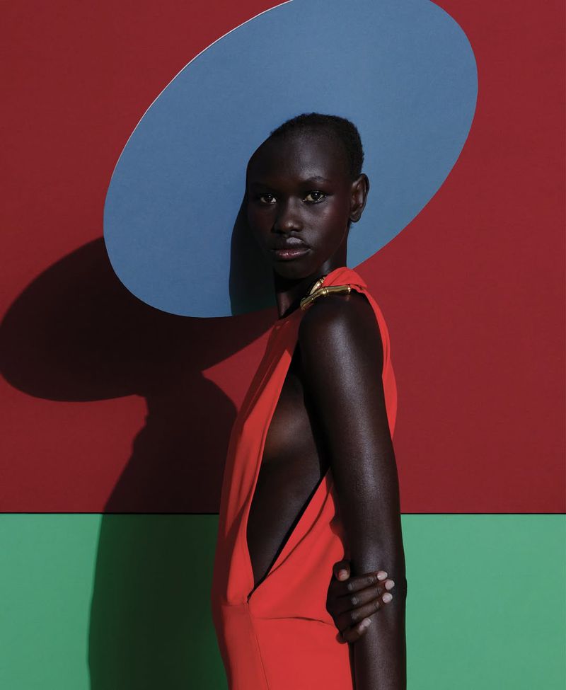Woman in a vibrant red dress and blue hat poses against a background with red and green blocks in an eye-level studio shot.