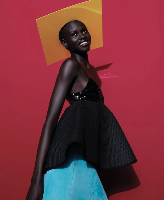 Portrait of a dark-skinned woman in a black top and blue skirt with a yellow square hat against a vibrant red background.