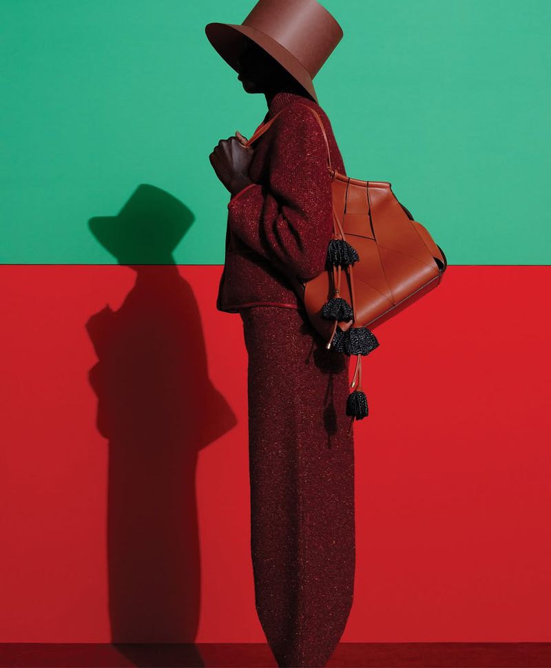 Dark-skinned woman wearing a tailored suit and hat stands in a studio with a red and green wall, carrying a leather purse.