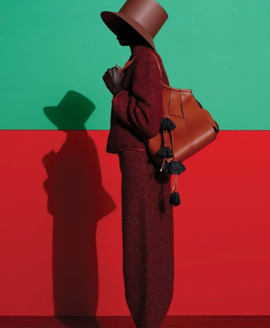 Dark-skinned woman wearing a tailored suit and hat stands in a studio with a red and green wall, carrying a leather purse.