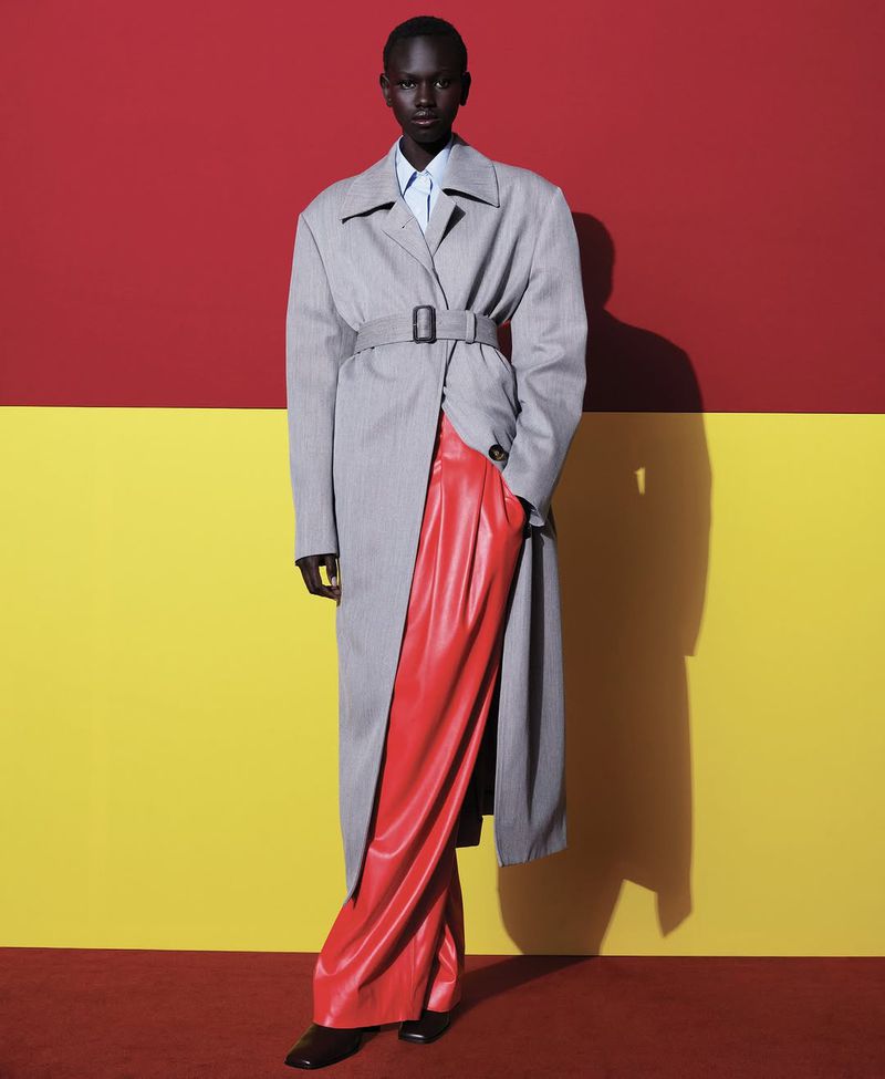 Editorial fashion shot of a Black female model in a grey belted trench coat, red pants, and black shoes, posing against a two-tone backdrop.
