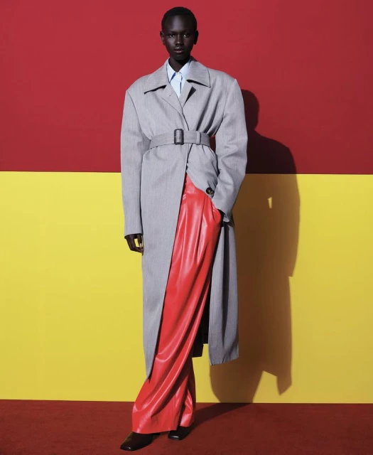 Editorial fashion shot of a Black female model in a grey belted trench coat, red pants, and black shoes, posing against a two-tone backdrop.