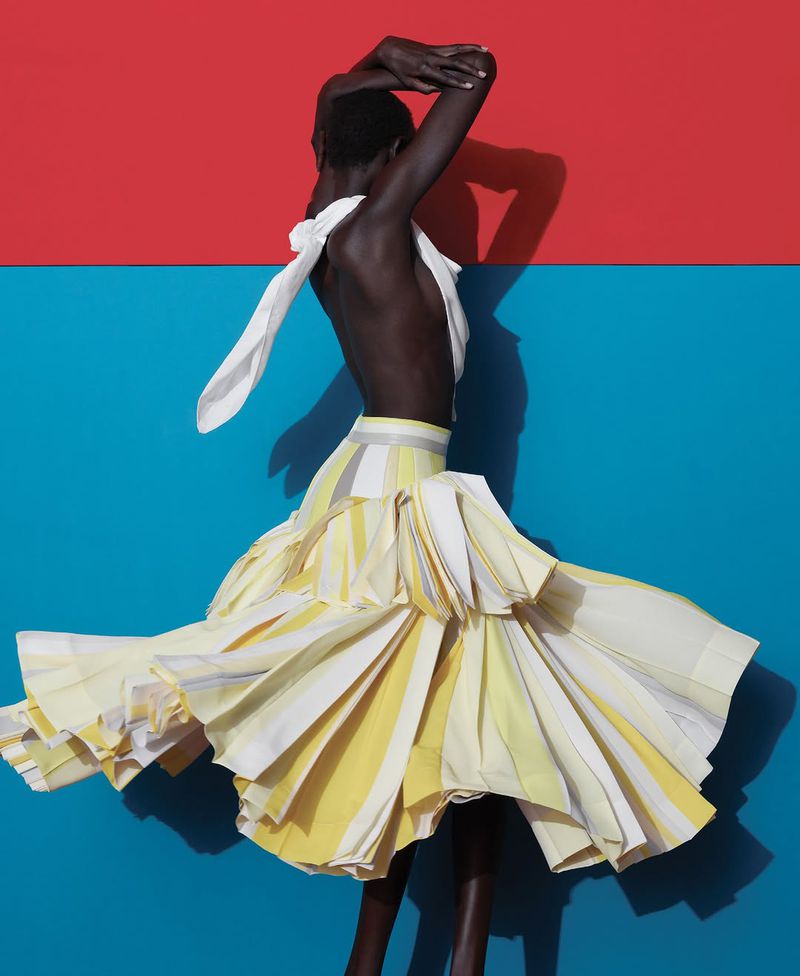 A dark-skinned model wears a layered, striped yellow and white skirt and white top against a split red and blue background.