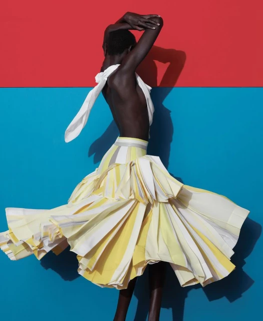 A dark-skinned model wears a layered, striped yellow and white skirt and white top against a split red and blue background.
