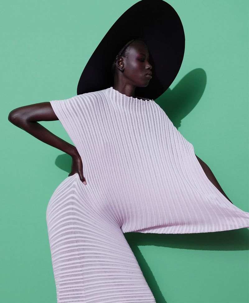 Dark-skinned woman poses in a light pink pleated outfit and large black hat against a green background in a fashion style photograph.