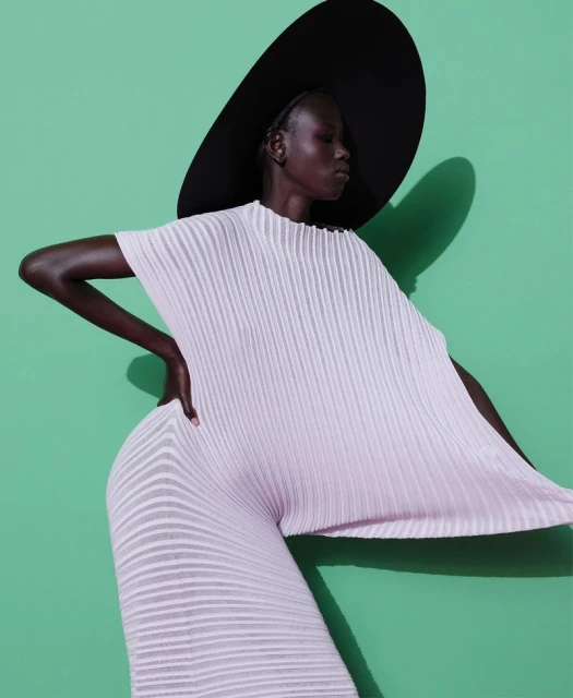 Dark-skinned woman poses in a light pink pleated outfit and large black hat against a green background in a fashion style photograph.