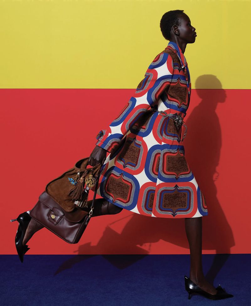 Fashion editorial shot of a Black female model in a patterned coat walking with a brown leather bag, set against a colorful backdrop.