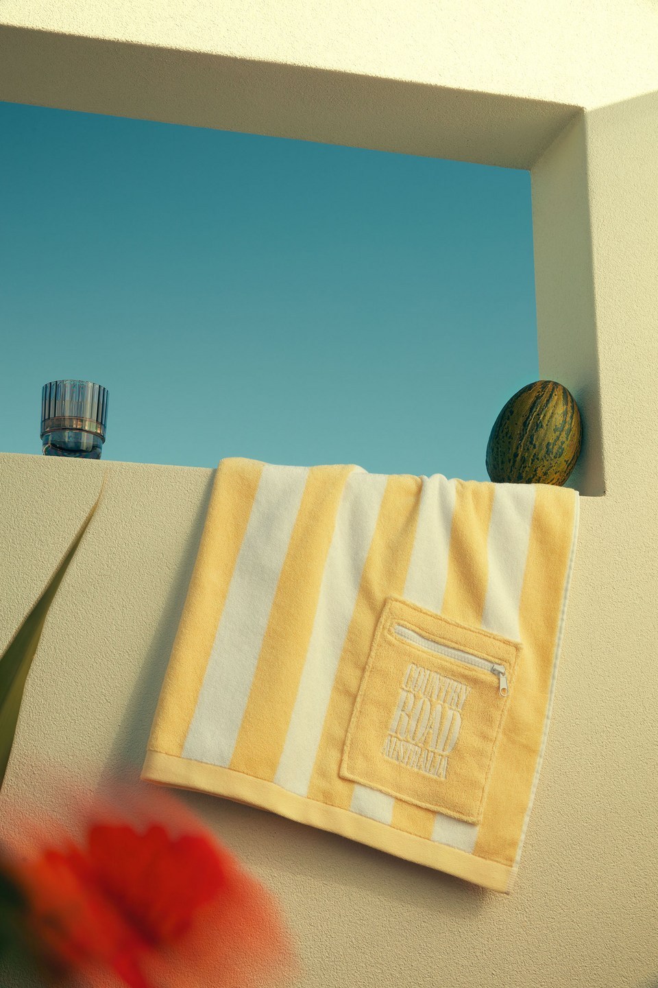 Still life featuring a yellow and white striped beach towel, a glass, and a melon, set against a bright blue sky.