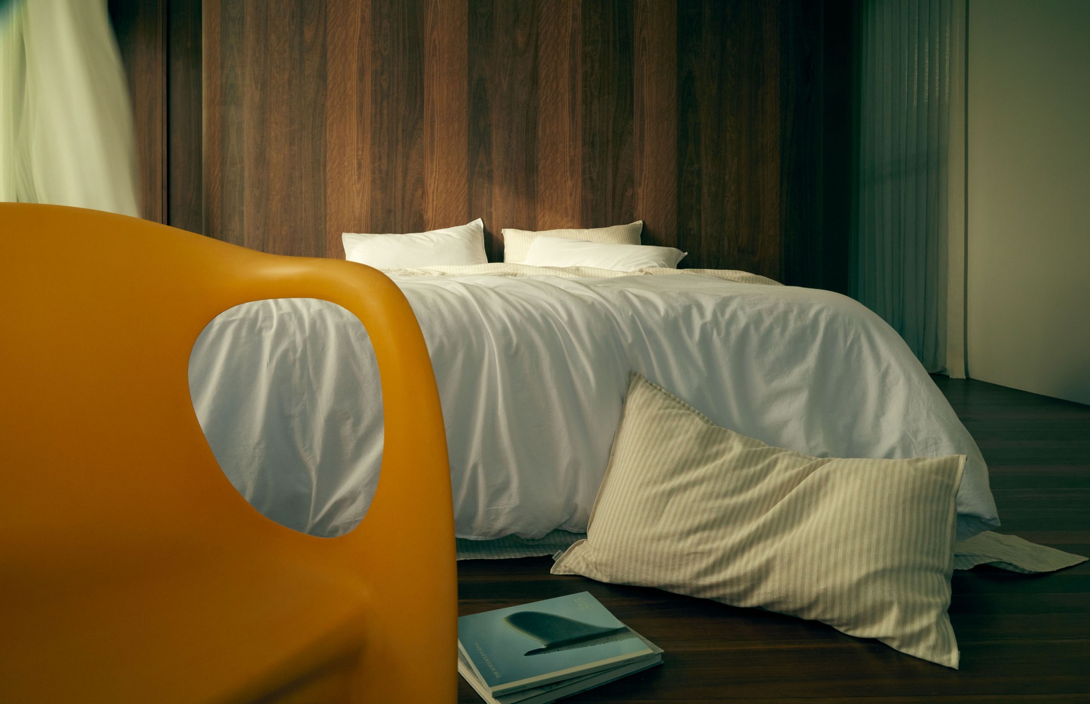 Minimalist bedroom interior featuring a white duvet bed, mustard chair, wooden headboard and wall, and natural light. 