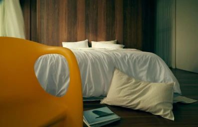 Interior bedroom view featuring a made white bed with wooden headboard and an orange chair in foreground