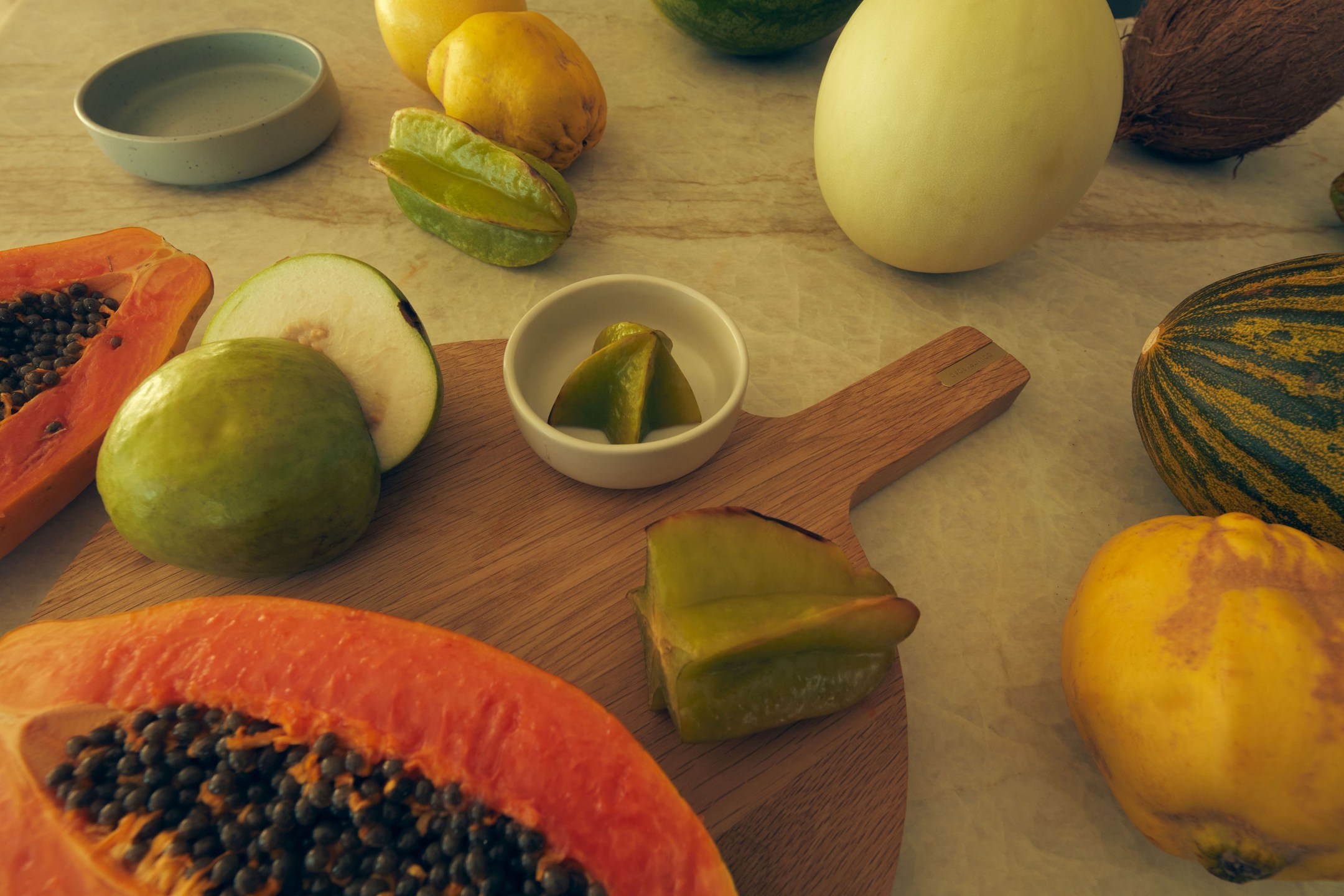 A variety of cut tropical fruits, including papaya, guava, and starfruit, are displayed on a wooden cutting board and marble surface.