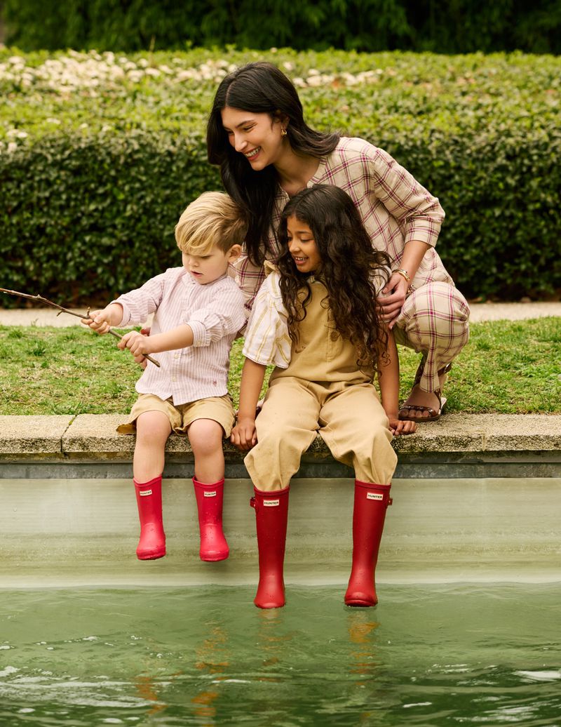 Mother and two children sitting by a pond wearing red boots. The mother is wearing a plaid outfit and smiling at the children.