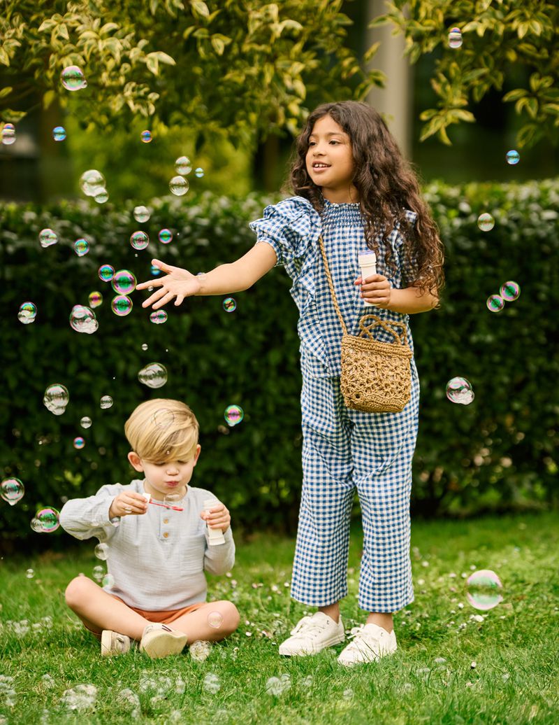 Two children, a girl and a boy, play with soap bubbles in a garden setting. The girl wears a blue gingham outfit, and the boy is seated on the grass.