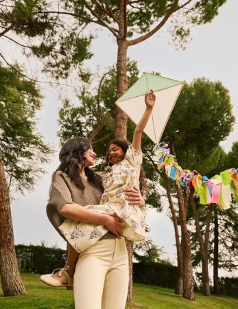 A happy family moment outdoors: a mother holding her young daughter, who is holding a kite, surrounded by green grass and trees.