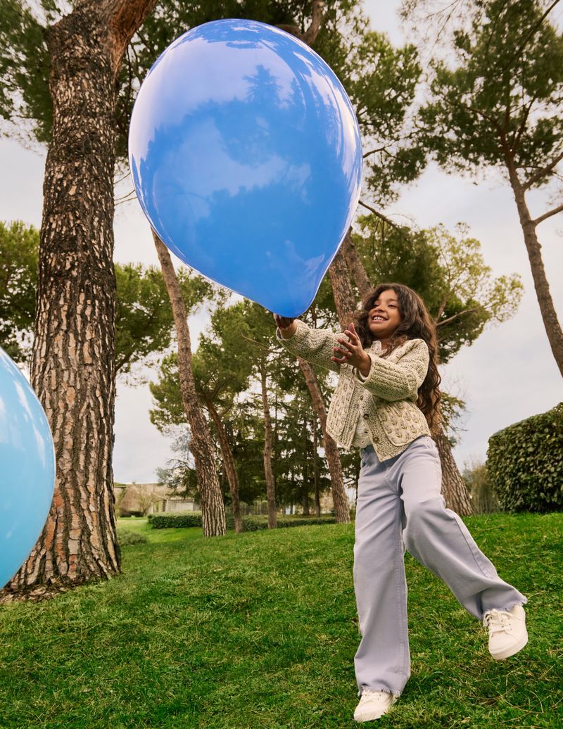 A young girl with long brown hair holds a large, bright blue balloon in a grassy park with tall trees, creating a joyful outdoor scene.