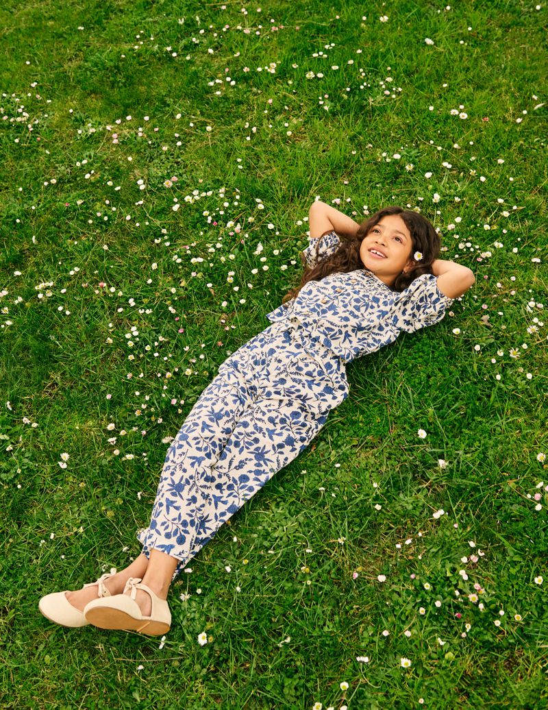 A child lies in grass with white flowers, wearing a blue and white floral print outfit and beige Mary Jane shoes, eyes looking up.