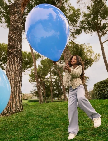 A young girl in a textured cardigan and light trousers reaches up to touch a large blue balloon in a park.