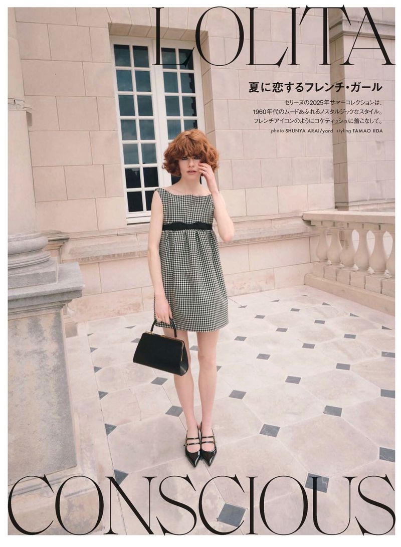 Fashion editorial image of a young adult female wearing a plaid dress, holding a black purse, posing on a stone terrace.