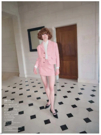 Fashion editorial shot of a woman in a pink skirt suit walking on a black-and-white checkered floor, with a door and artwork in the background.