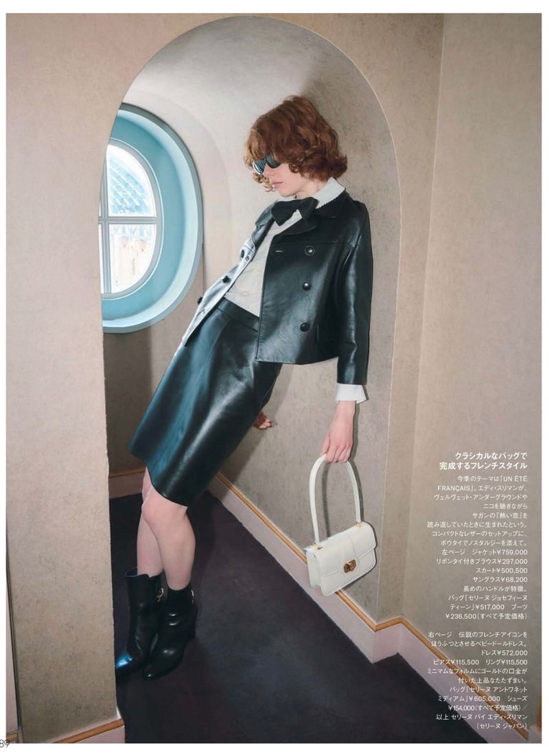 A red-haired woman in a black leather skirt suit and boots leans against an arched doorway, holding a white purse in a studio fashion shot.