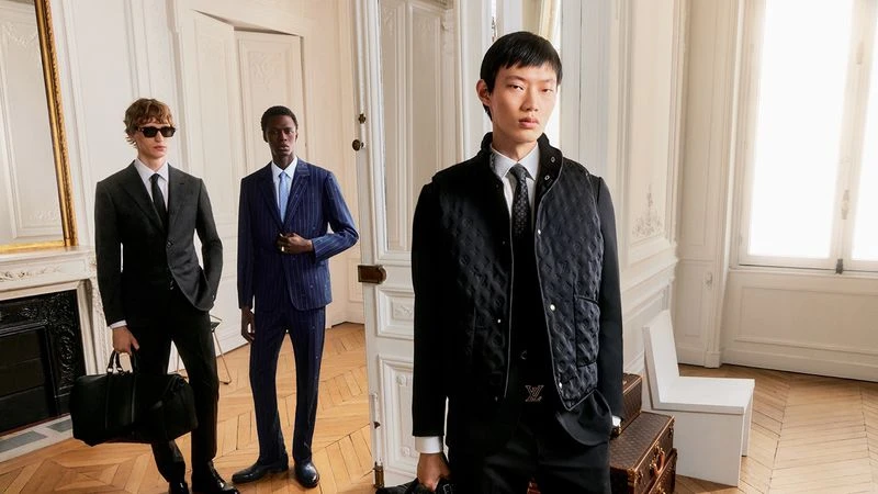 Three adult men in stylish suits and vests posing indoors against white walls and wood floors, showcasing high-end fashion.