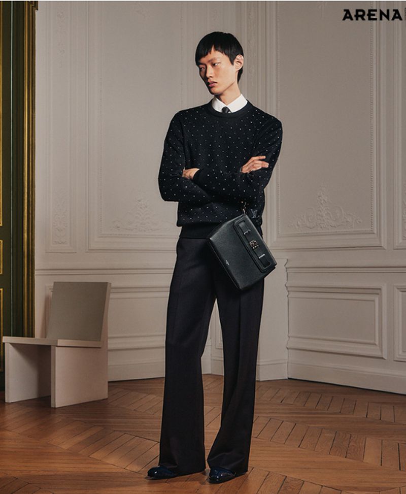 An Asian male model poses in a black sweater, trousers, and black leather messenger bag against an ornate wall and wooden floor.
