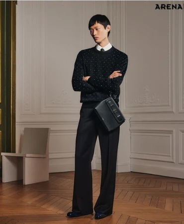 An Asian male model poses in a black sweater, trousers, and black leather messenger bag against an ornate wall and wooden floor.