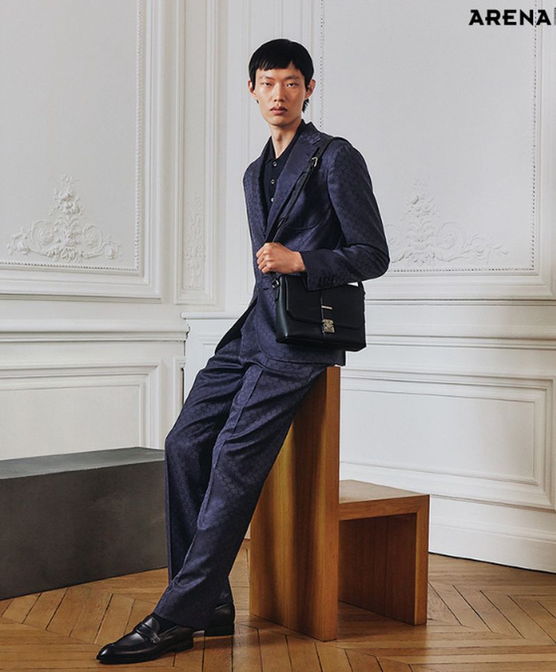 Asian man in a patterned suit poses indoors with a leather bag. He sits on a chair and is photographed against a decorative wall.
