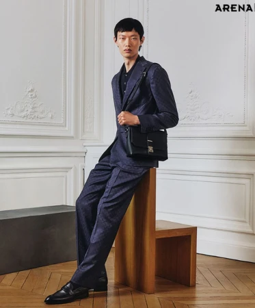 Asian man in a patterned suit poses indoors with a leather bag. He sits on a chair and is photographed against a decorative wall.