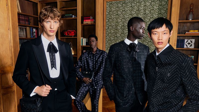 Four diverse men in formal attire pose indoors with a bookcase, showcasing high-end fashion in a sophisticated and elegant setting.