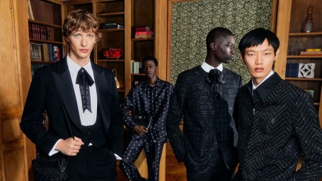 Four male models in dark tailored suits and patterned apparel standing in a wood-paneled library interior for Louis Vuitton.