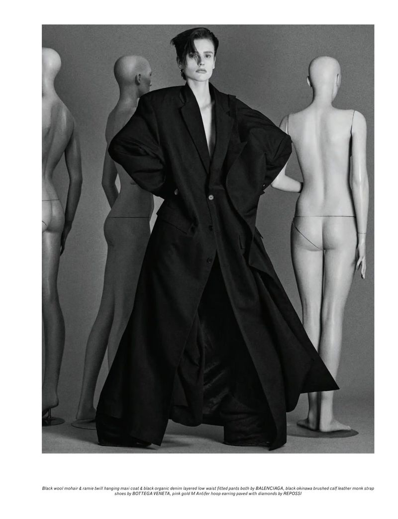 Editorial fashion shot of a model wearing an oversized black coat and pants, posing in front of nude-colored mannequin torsos.