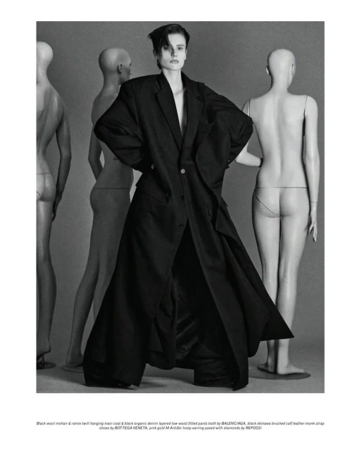 Editorial fashion shot of a model wearing an oversized black coat and pants, posing in front of nude-colored mannequin torsos.