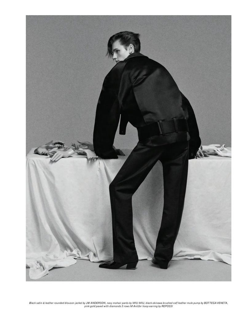 Model in satin jacket and trousers leaning on draped table in black and white fashion shot with artful styling in a studio setting.