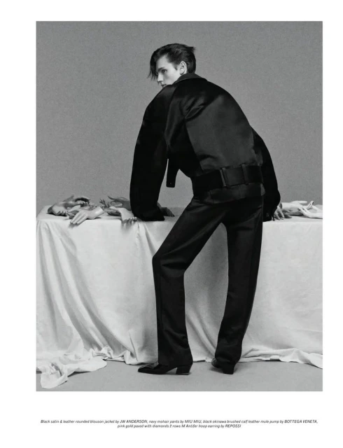 Model in satin jacket and trousers leaning on draped table in black and white fashion shot with artful styling in a studio setting.