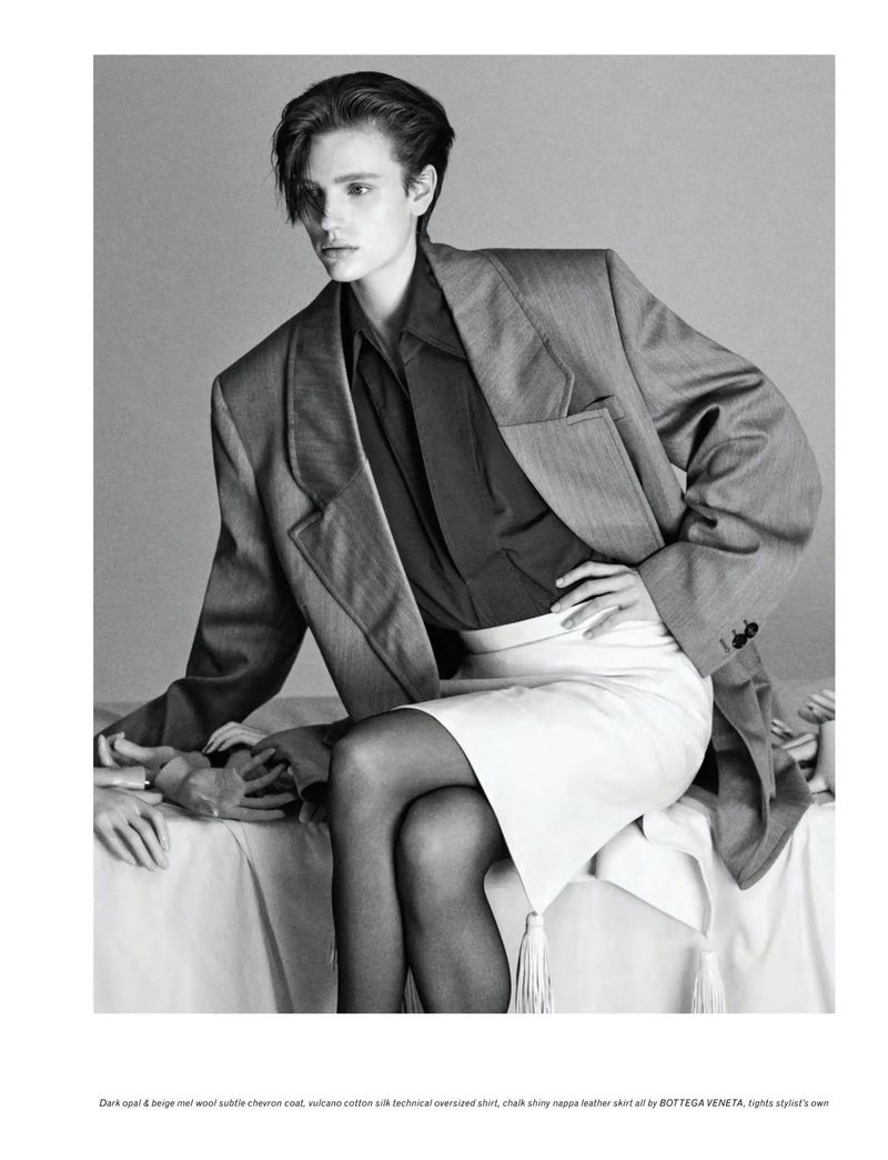 Fashion editorial shot: an androgynous model in an oversized grey blazer and white skirt sits with their hand resting on a person's hand.